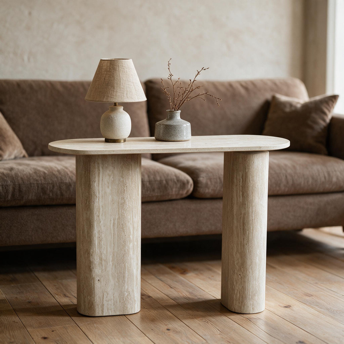 Smalle sidetable in travertijnlook op twee ovale zuilen, achter een zetel met lamp en vaas